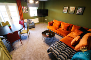 Set in the green living room of our accommodation in Huntingdon, an orange velvet corner sofa opens to the living room where colourful chairs are set around a circular dining table.