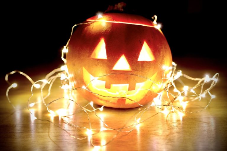 A smily-face pumpkin is decorated with fairy lights against a dark background.